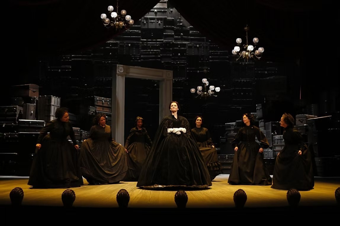 A production photo of women in black mourning-era costumes on a stage set built from stacked steamer trunks and crates